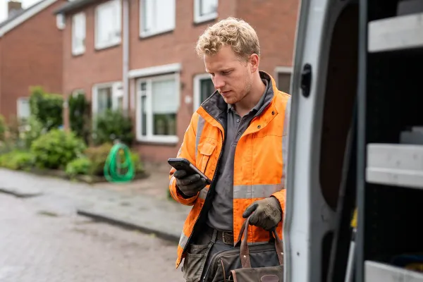Een monteur in werkjas kijkt op zijn telefoon bij de open zijdeur van een bestelbus.