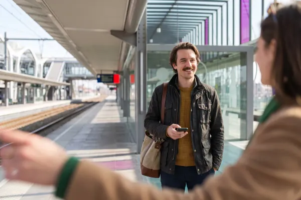 Een man met zijn telefoon in de hand lacht naar een vriendin op het perron van een station.