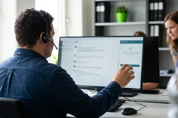 Een servicedeskmedewerker met headset bekijkt een voorstel op zijn scherm terwijl een collega meekijkt.