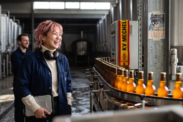 Een productieleider met een tablet lacht om een grap van een collega naast de bottellijn van een brouwerij.