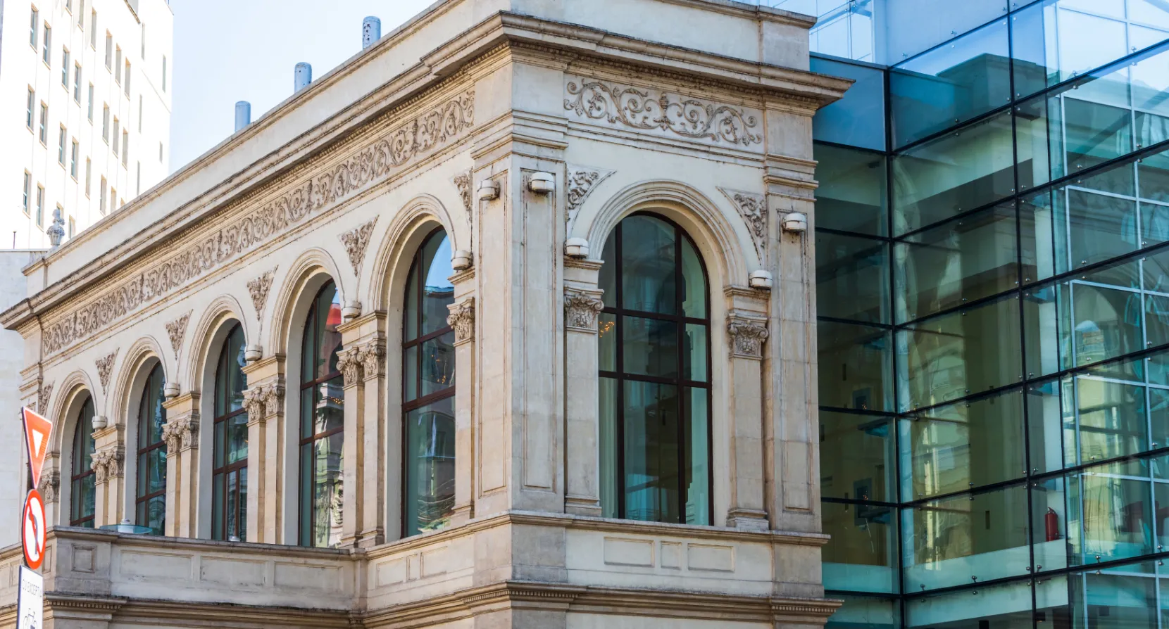 Een klassiek bankgebouw met rijk versierde gevel, naast een moderne glazen aanbouw.