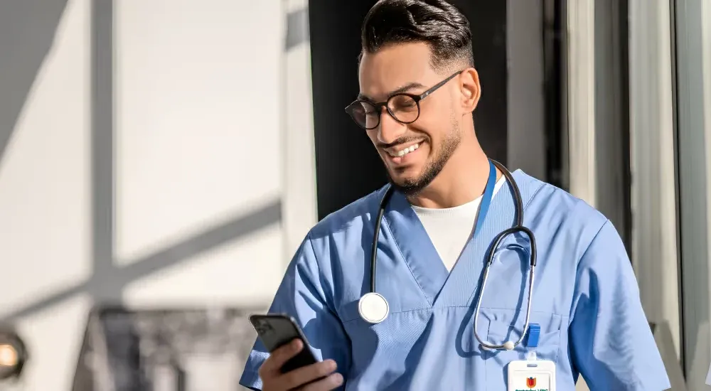 Een lachende verpleegkundige in een blauw uniform kijkt op zijn smartphone.